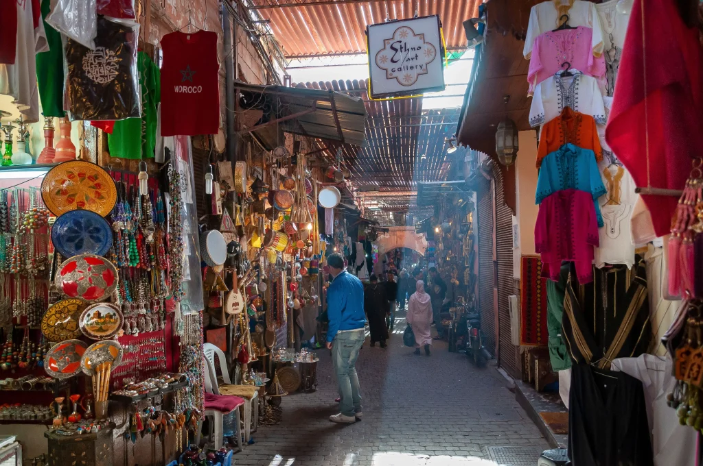 A narrow Moroccan market alley lined with shops displaying metal plates, jewelry, and colorful clothing, with a man walking through the center.