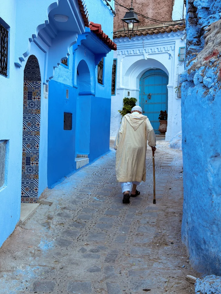 Elderly man in traditional robe walking in a sunlit Moroccan alleyway.
